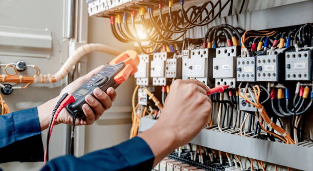 Electrician in a cap, yellow glasses, and an orange vest uses a digital multimeter to test electrical current on a distribution box for an EICR report.