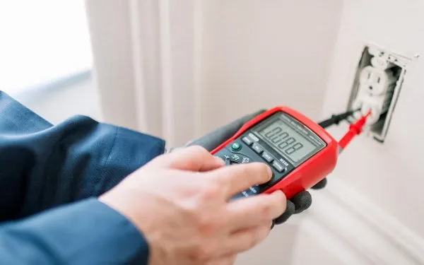 A person wearing gloves uses a digital multimeter to test the voltage of a wall electrical socket, with the display showing a reading and probes inserted into the socket.