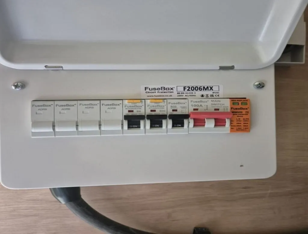 A white consumer unit with labelled switches, including several circuit breakers and a main switch, mounted on a light wood surface. The cover is open, revealing the labelled controls inside.