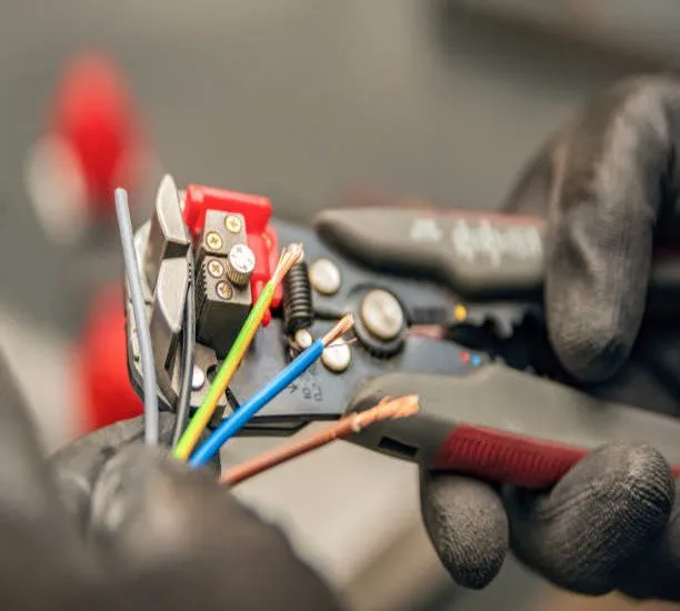 A person wearing black gloves uses a wire stripping tool to strip insulation from several coloured electrical wires, exposing the copper conductors.