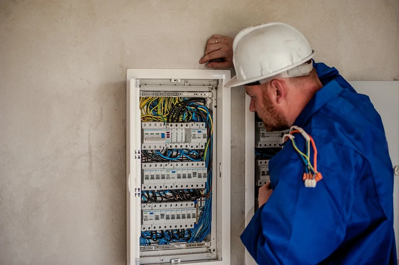 An electrician wearing a white hard hat and blue work clothes is inspecting an open electrical panel with many coloured wires and switches on a beige wall.