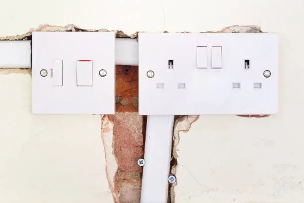 Two wall-mounted white electrical sockets and switches are shown; the plaster around them is broken, revealing red bricks and wiring beneath the surface—ideal for inspection by a certified rewiring electrician.