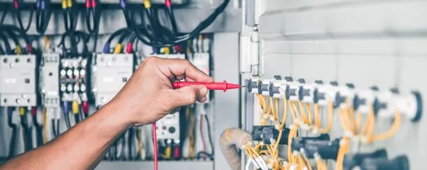 A person’s hand uses a red electrical tester probe on terminal connections inside an electrical control panel filled with wires and components.