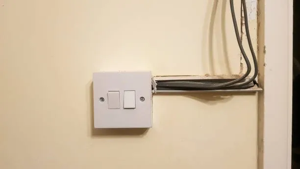 A white electrical light switch with exposed wires and an unfinished opening in the wall, highlighting untidy installation—ideal for a certified rewiring electrician or house rewiring Tooting project.