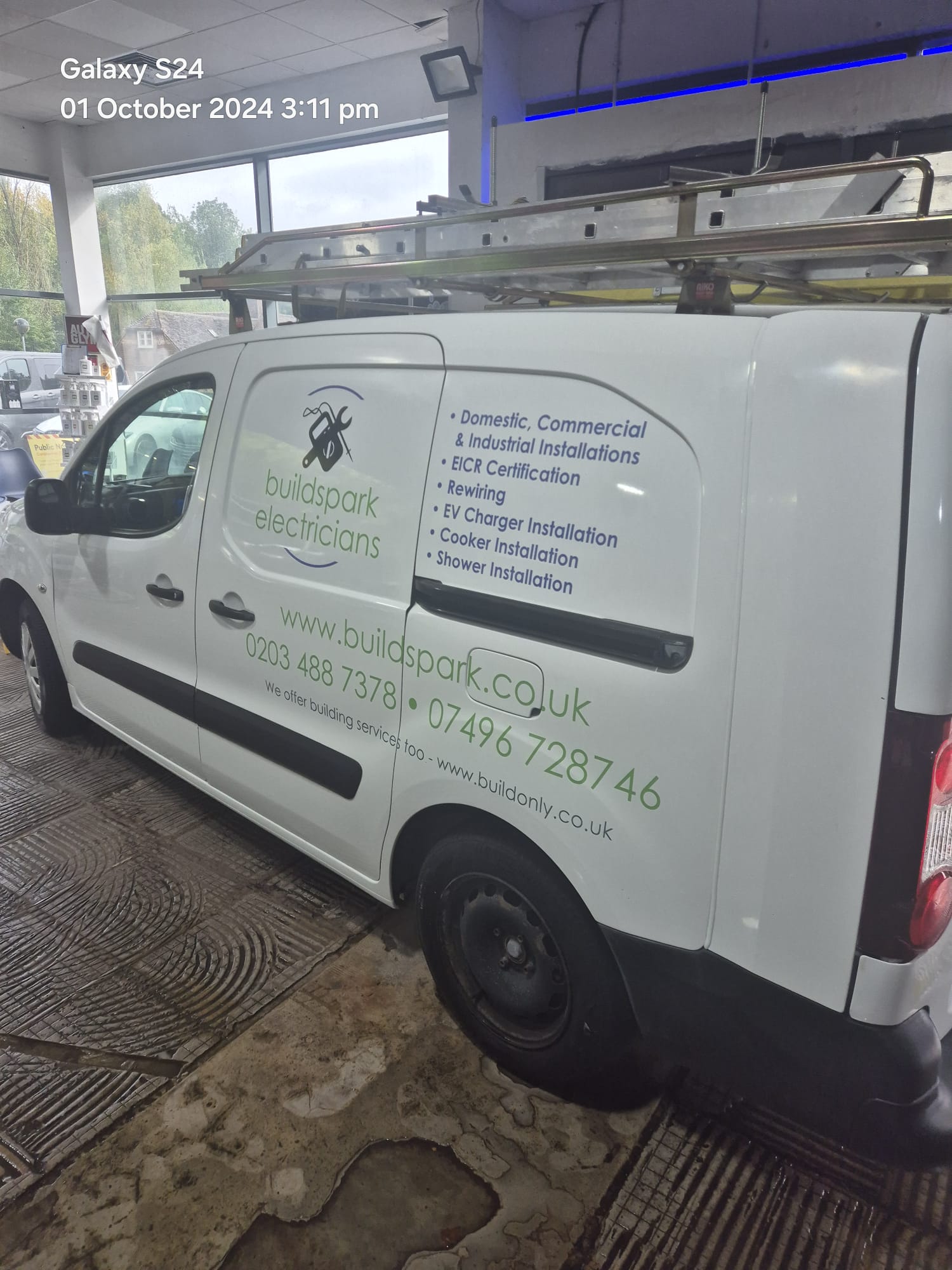 A white van with Build Spark Electricians branding and contact info is parked indoors. A ladder is mounted on the roof, and the van advertises services like EV charger installation and electrical certification.