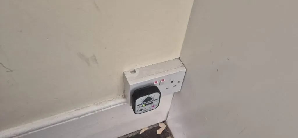 A double power socket on a wall, with a black electrical device plugged into the left socket. The socket has two red switches and is mounted near a scuffed wall and skirting board. Peeling paint is visible on the floor below.