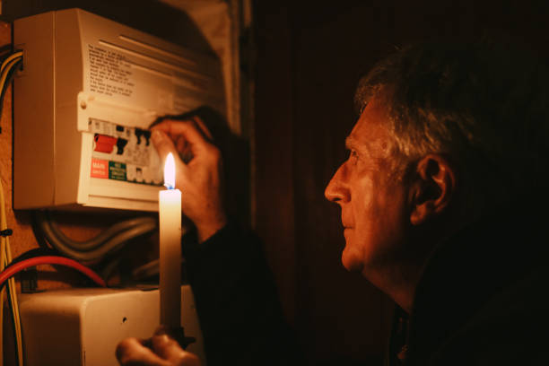 An older man holds a lit candle while inspecting and adjusting switches on an electrical fuse box during a power cut, awaiting urgent electrical repairs in Wimbledon. The scene is dimly lit by the candlelight.